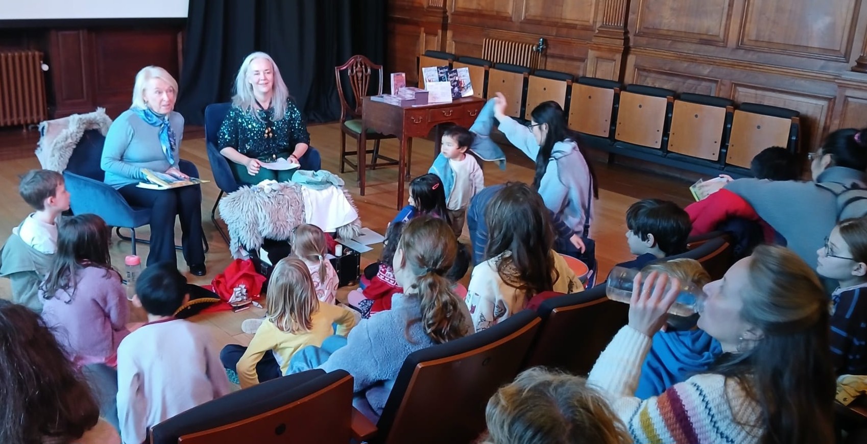 A beautiful wood-panelled room with children of a range of ages seated, some with parents, in stands rising towards the photographer. Authors,Paula F Andrews and Maura McRobbie are seated facing the audience.